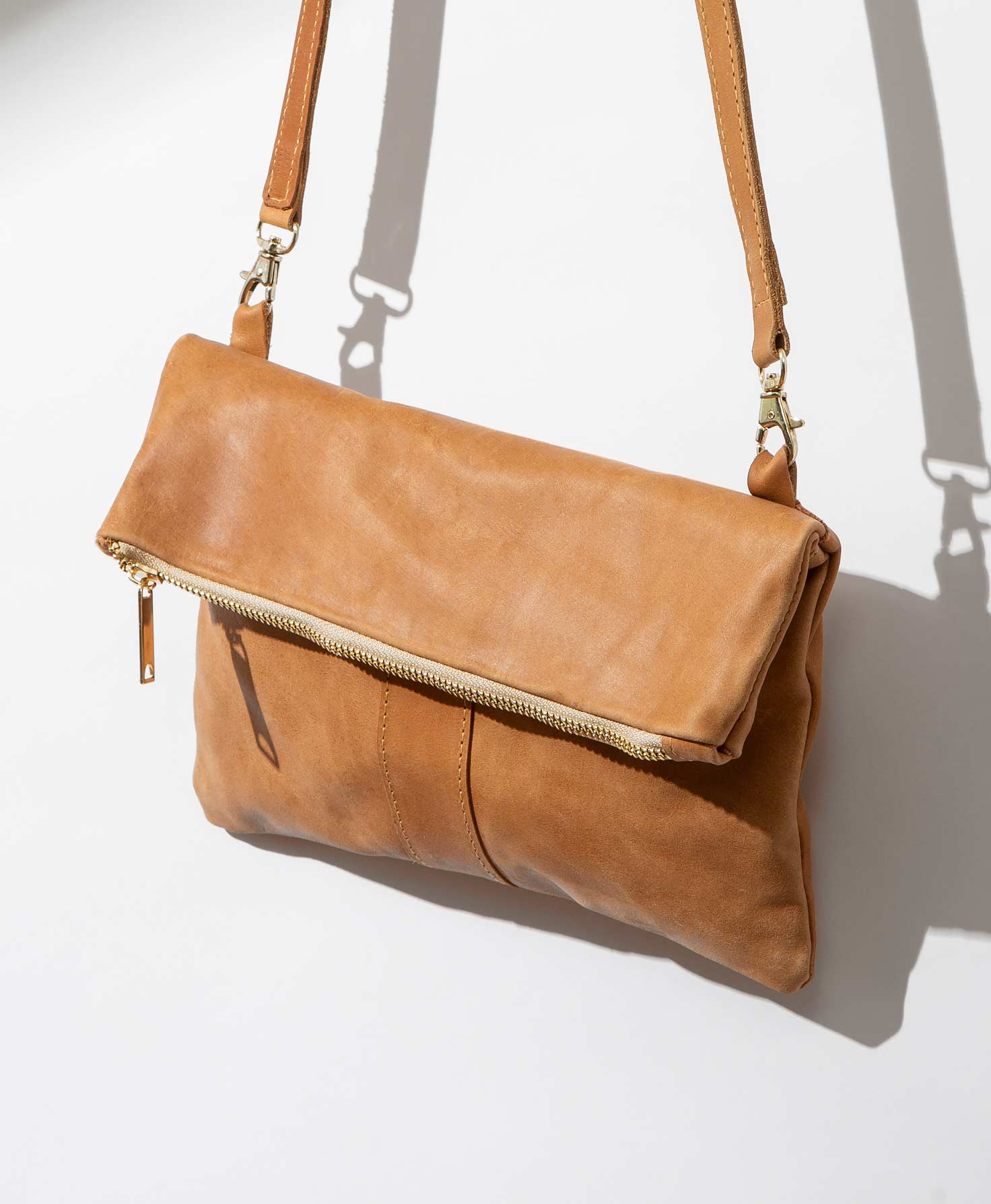 The Adwa Foldover Clutch is shown hanging against a white wall. It is made of buttery-soft, caramel-colored leather. The top third of the bag is folded over, and to open the bag the wearer unfolds it and unzips the golden zipper at the top. The straps are connected to the top of the fold via two golden clips positioned on either side of the bag. The long leather strap can be adjusted or removed entirely. 