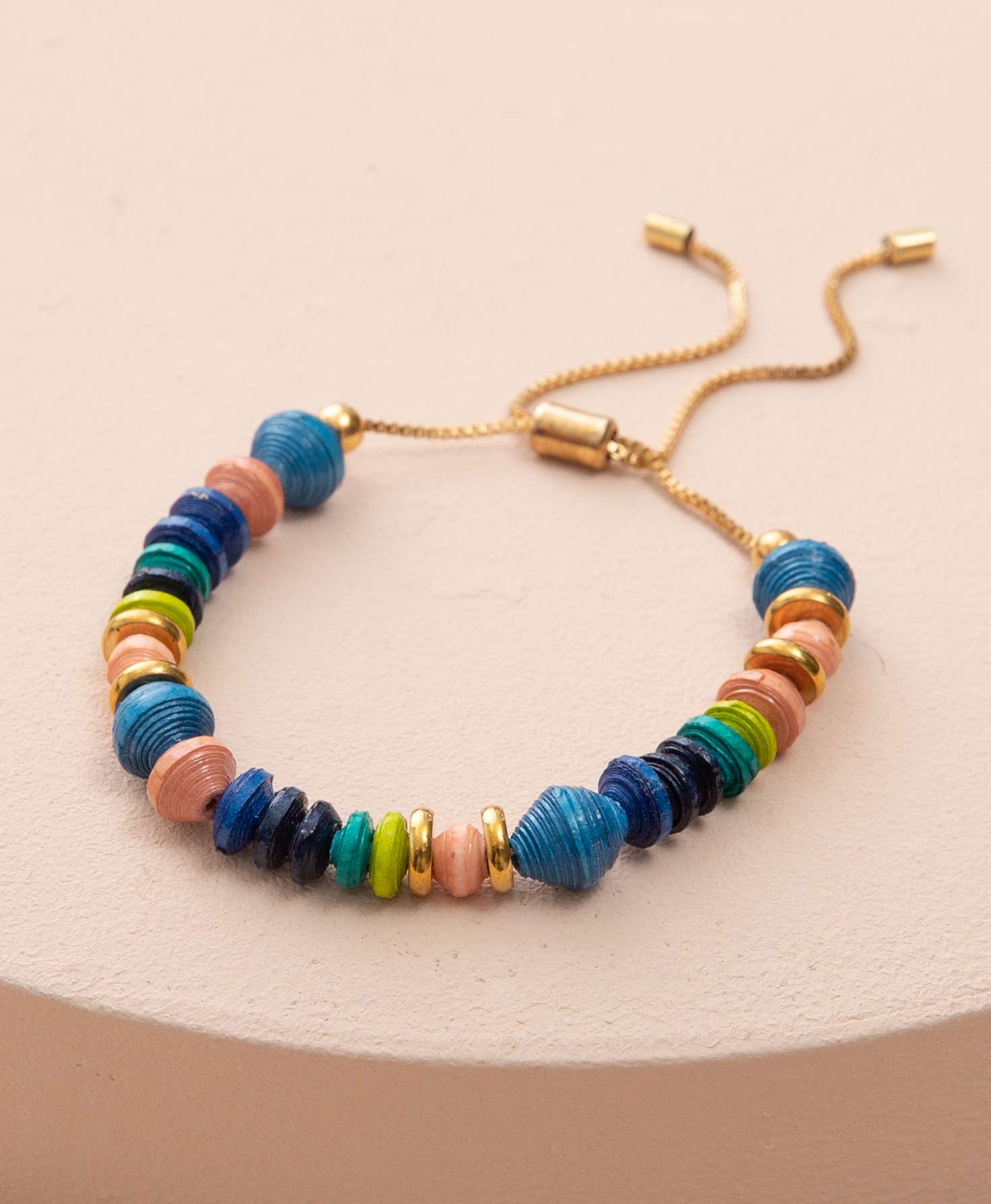 The Bevy Bracelet lays on a peach-colored block. Hand-rolled paper beads in alternating shades of blush, blue, navy, turquoise, and lime green are strung on a dainty gold chain. The paper beads have a variety of shapes. Some are disc-shaped while others are round. The bracelet is accented with a few gold metal discs. The closure of the bracelet is adjustable and can be tightened by pulling the two ends of the chain in opposite directions. 