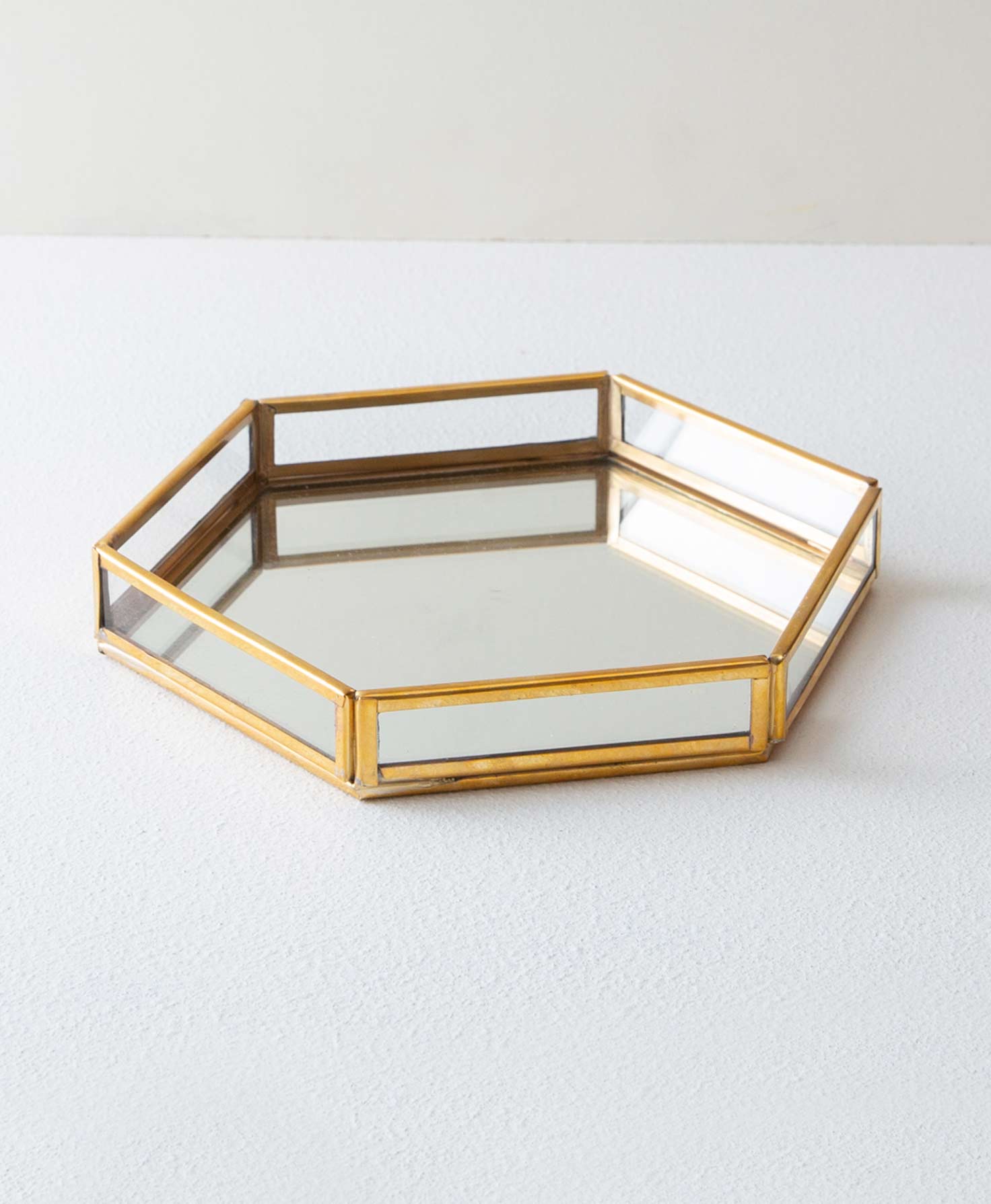 The Brass and Glass Hexagonal Tray sits on a white surface. It has six low sides composed of a rectangular glass panels framed in shining brass. The base of the tray is a hexagon made of mirrored glass.