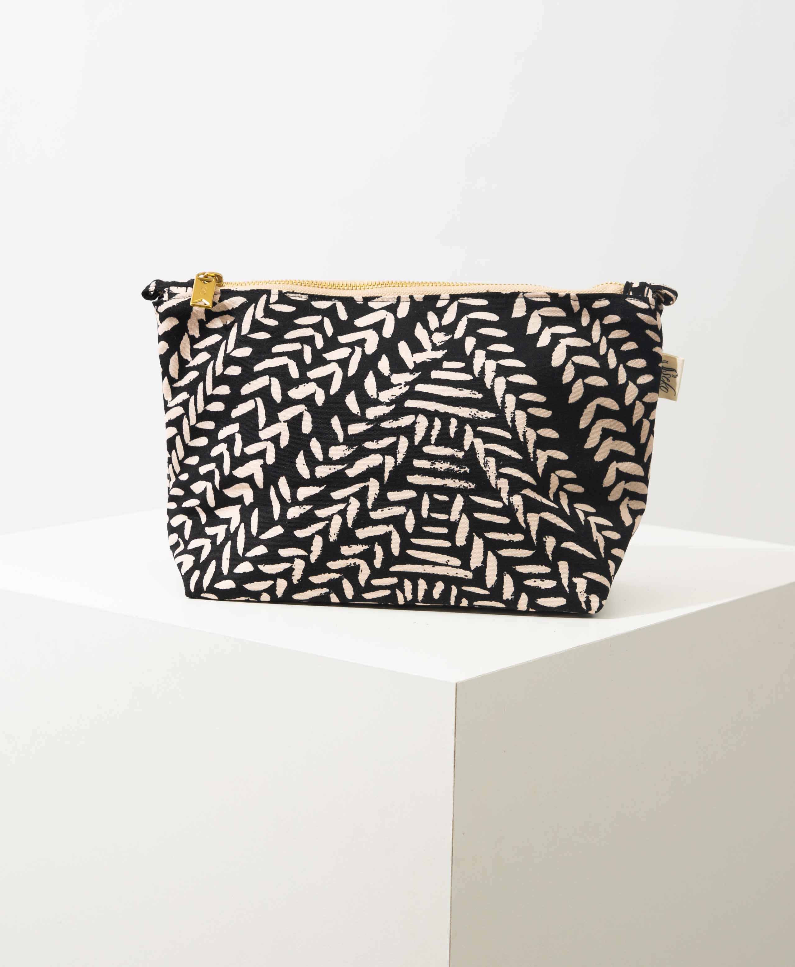 The Carryall Cosmetic Bag in Kawa print sits upright on a white block. It is roughly rectangular and is wider than it is tall. It is made of printed cotton that has a black background with white, hand-painted looking markings. There is a brass zipper along the top of the bag, and two fabric loops on either corner, allowing the bag to turn into a crossbody. 