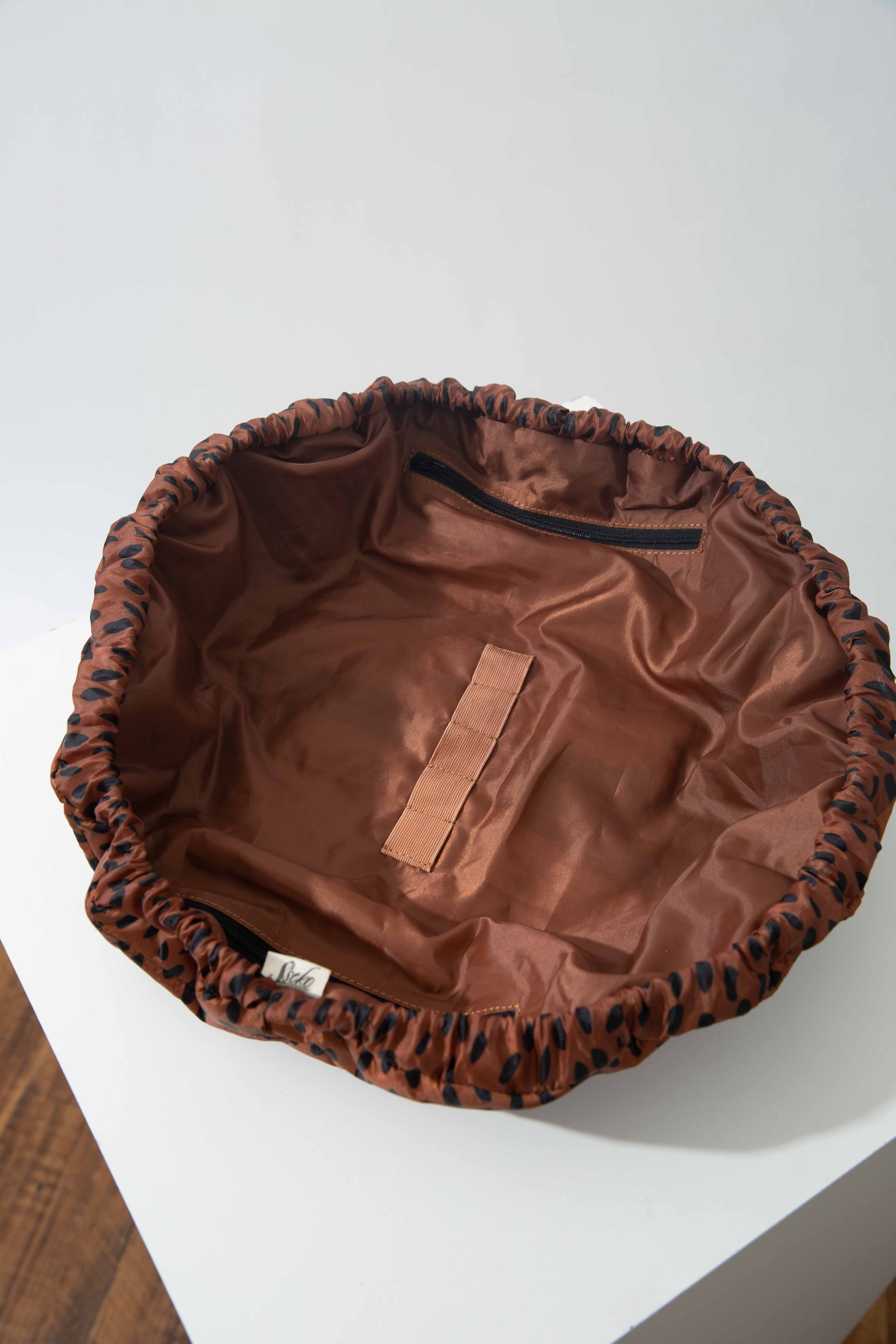 The Catchall Drawstring Pouch sits on a white block and is shown from overhead. It is fully opened, and has a wide round mouth. The interior lining of the pouch is a brown satin. There is a zip pocket stitched into the side of the interior, and there are five small elastic loops stitched to the bottom to hold items in place. 