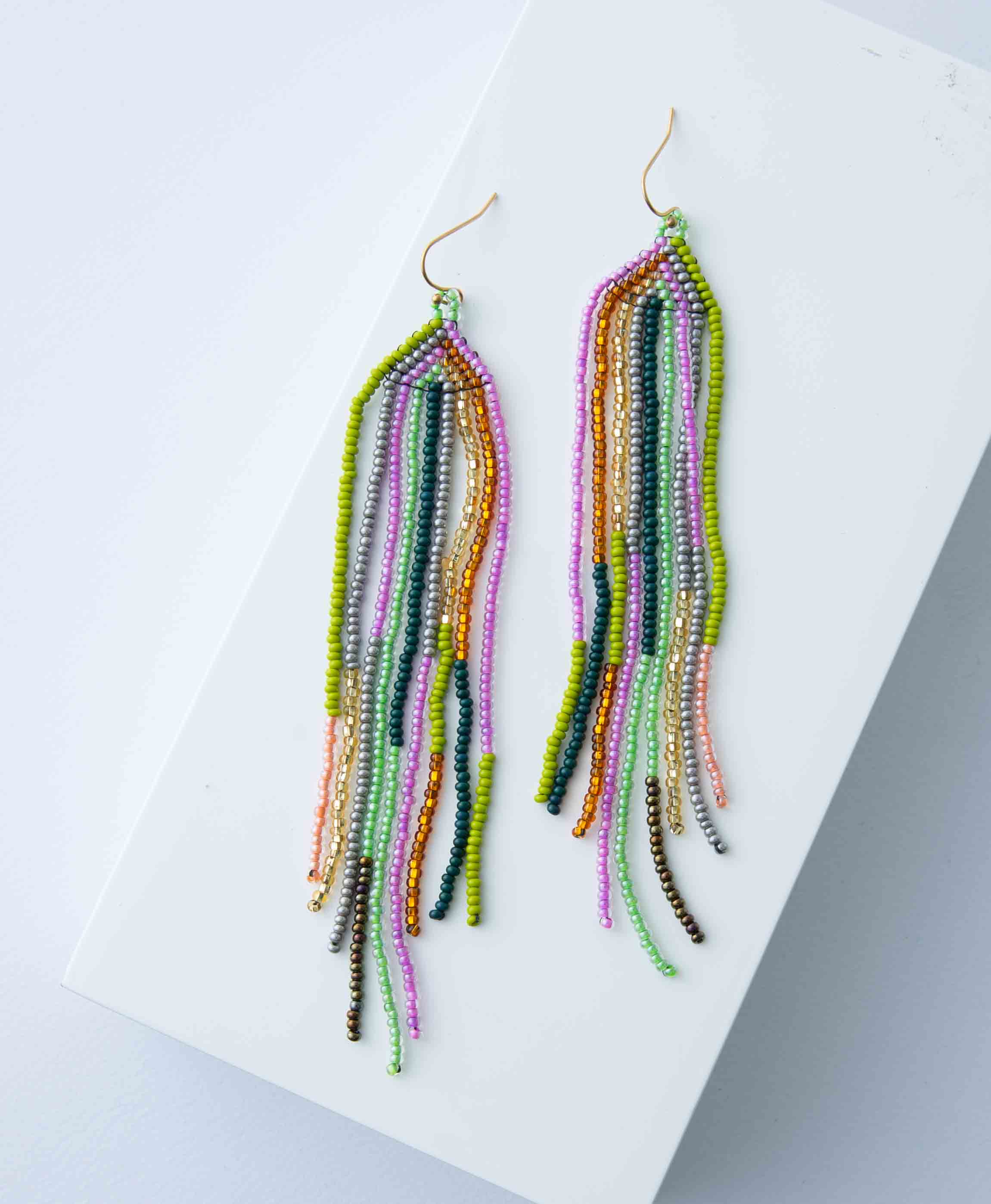 The Colores Earrings lay against a white background. They are long chandelier-style earrings composed of many strands of dainty glass beads. The strands are various lengths and include beads in shades of mint, pink, gold, navy, green, bronze, and silver. 