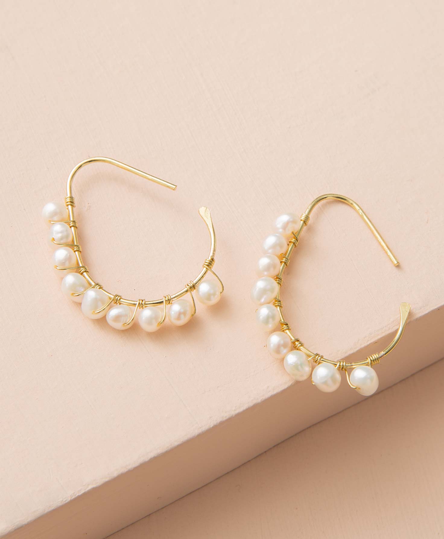 The League Earrings lay flat on a peach colored background. A gold wire forms the base of the earrings, and is looped around to form an oblong hoop shape. Attached along the hoop via gold wire is a row of 9 genuine pearls. The pearls vary slightly in size and shape and have a natural feel. 