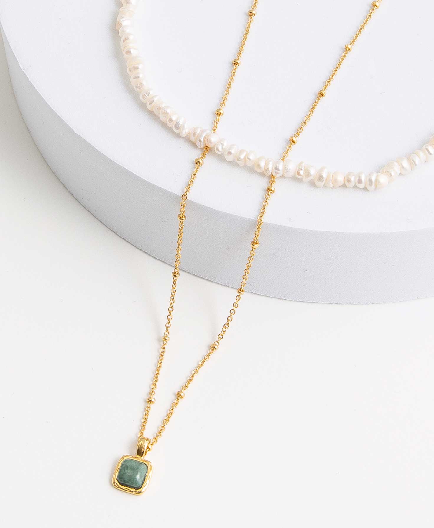 The two necklaces included in the Luster Necklace set lay draped on a white block. The shorter necklace is composed entirely of a string of shining pearls with a natural, imperfect shape. The longer necklace is a gold chain necklace with small brass beads spaced out along the length of the chain. At the bottom is a small pendant with a square of smooth, forest green marble outlined in brass.