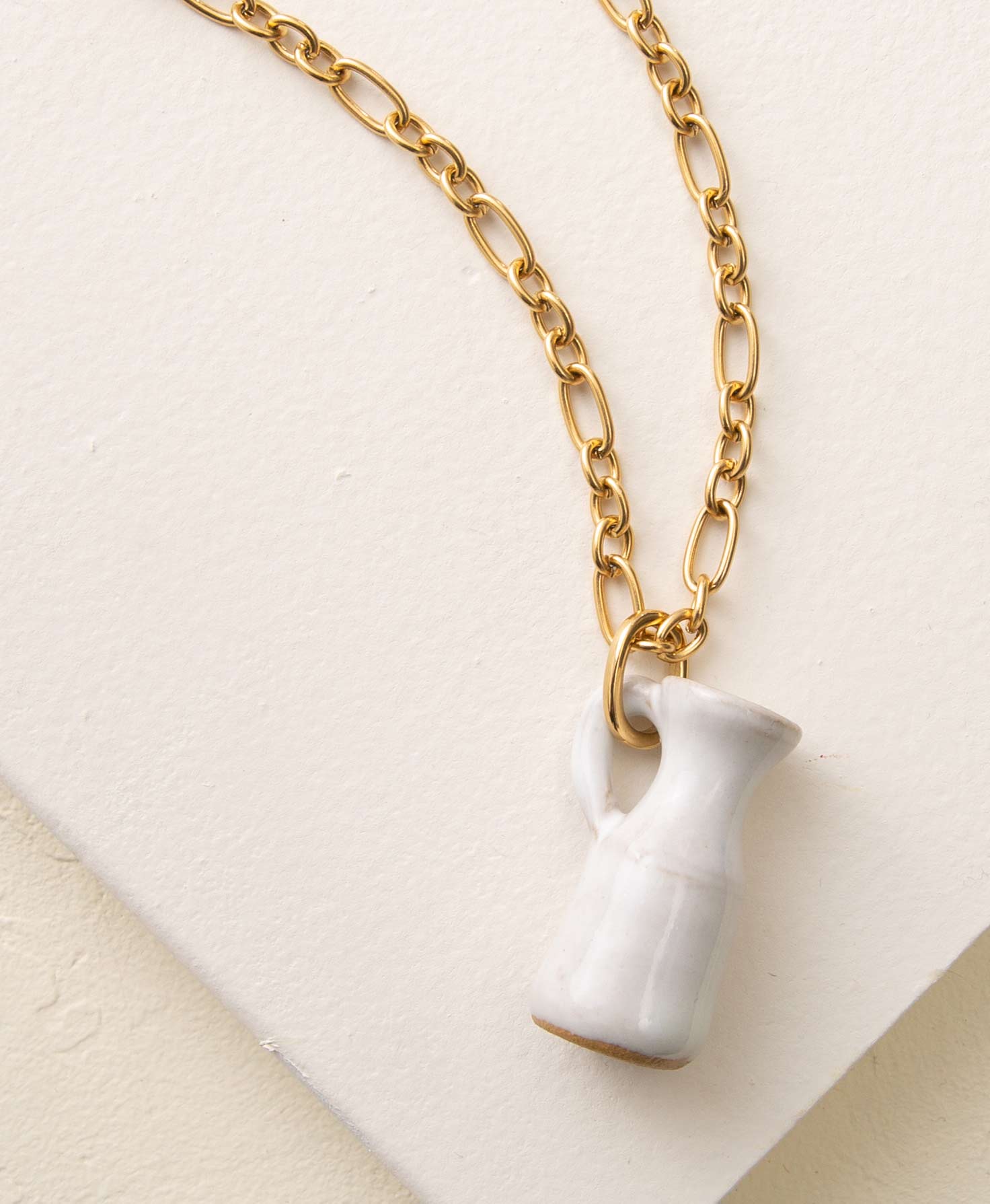 The Petite Pitcher Necklace lays on a white background. It has a chunky gold chain composed of circular and oval-shaped links. At the bottom of the chain is a white porcelain pendant shaped like a rustic pitcher. 