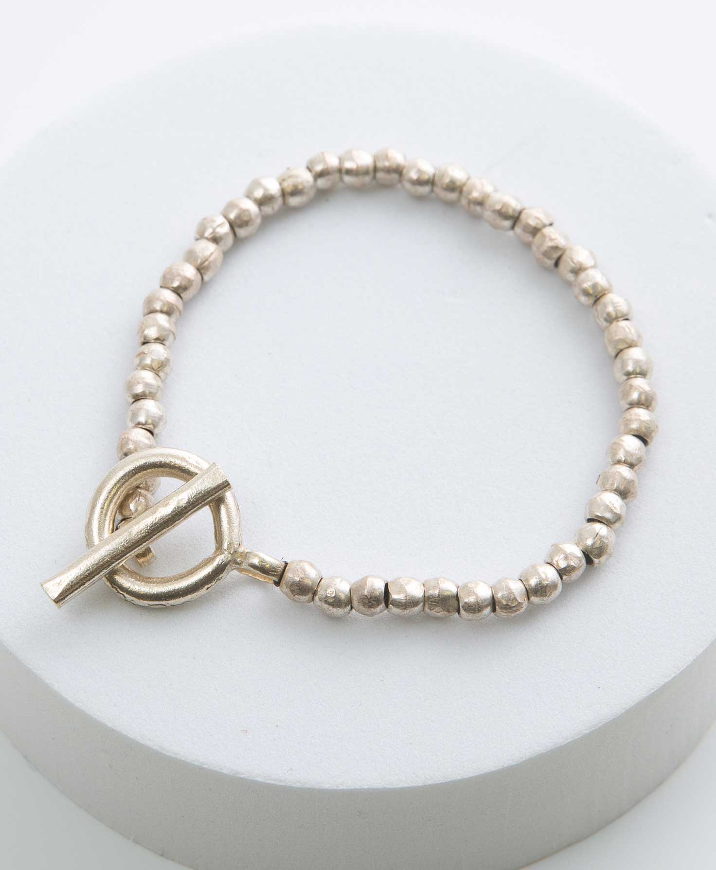 The Pristine Bracelet is clasped and sits on a white block. It is made of a single strand of round, silver beads with slightly irregular textures for a handmade look. The toggle clasp is also a design feature, and consists of a silver bar that rests within a silver loop.
