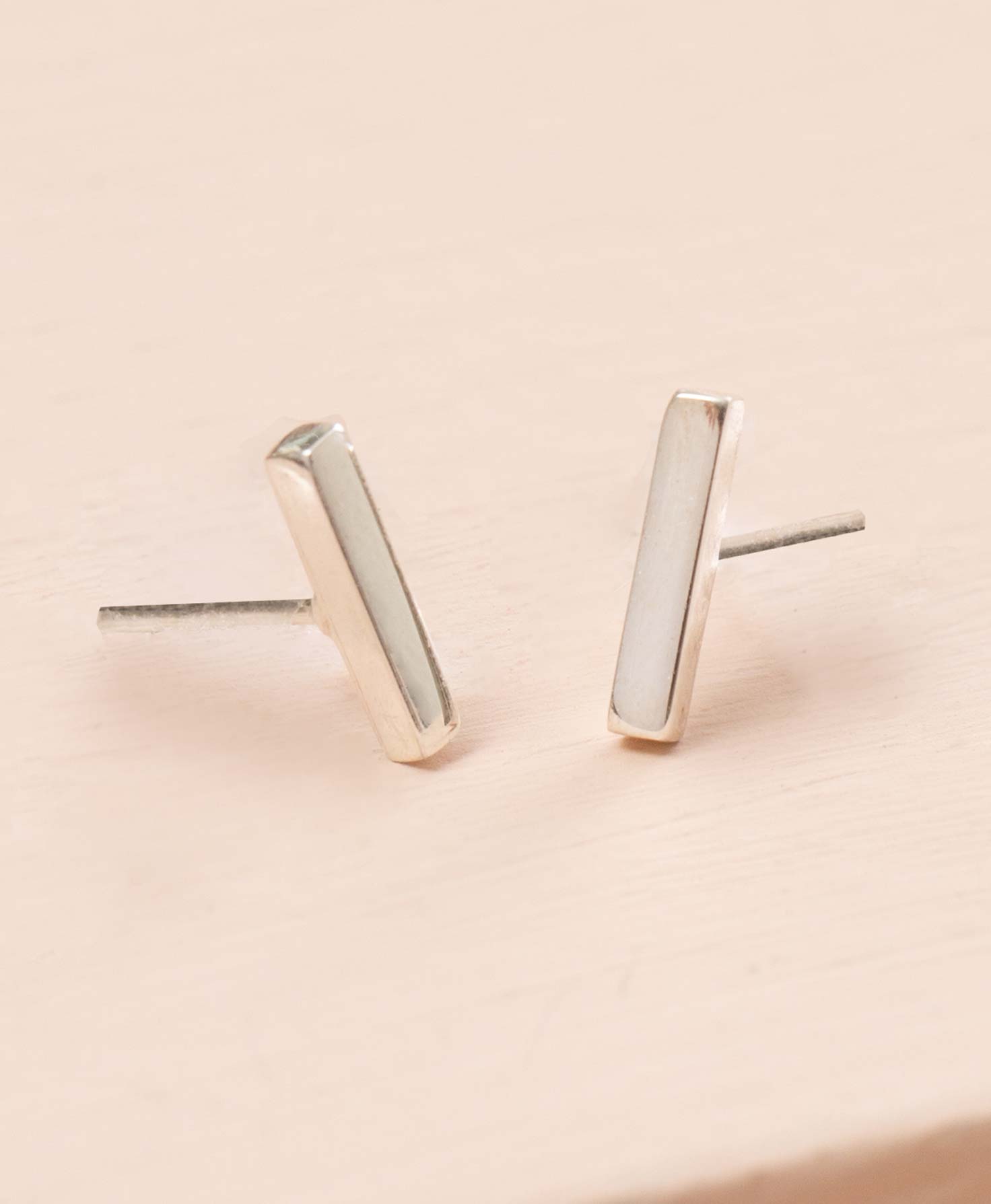 The Silver Bar Studs sit on a cream colored background. They are small, minimalistic studs made of silver plated brass. The silver posts are connected to a rectangular, 3-dimensional silver bar. The dainty bar has a shiny metallic finish.