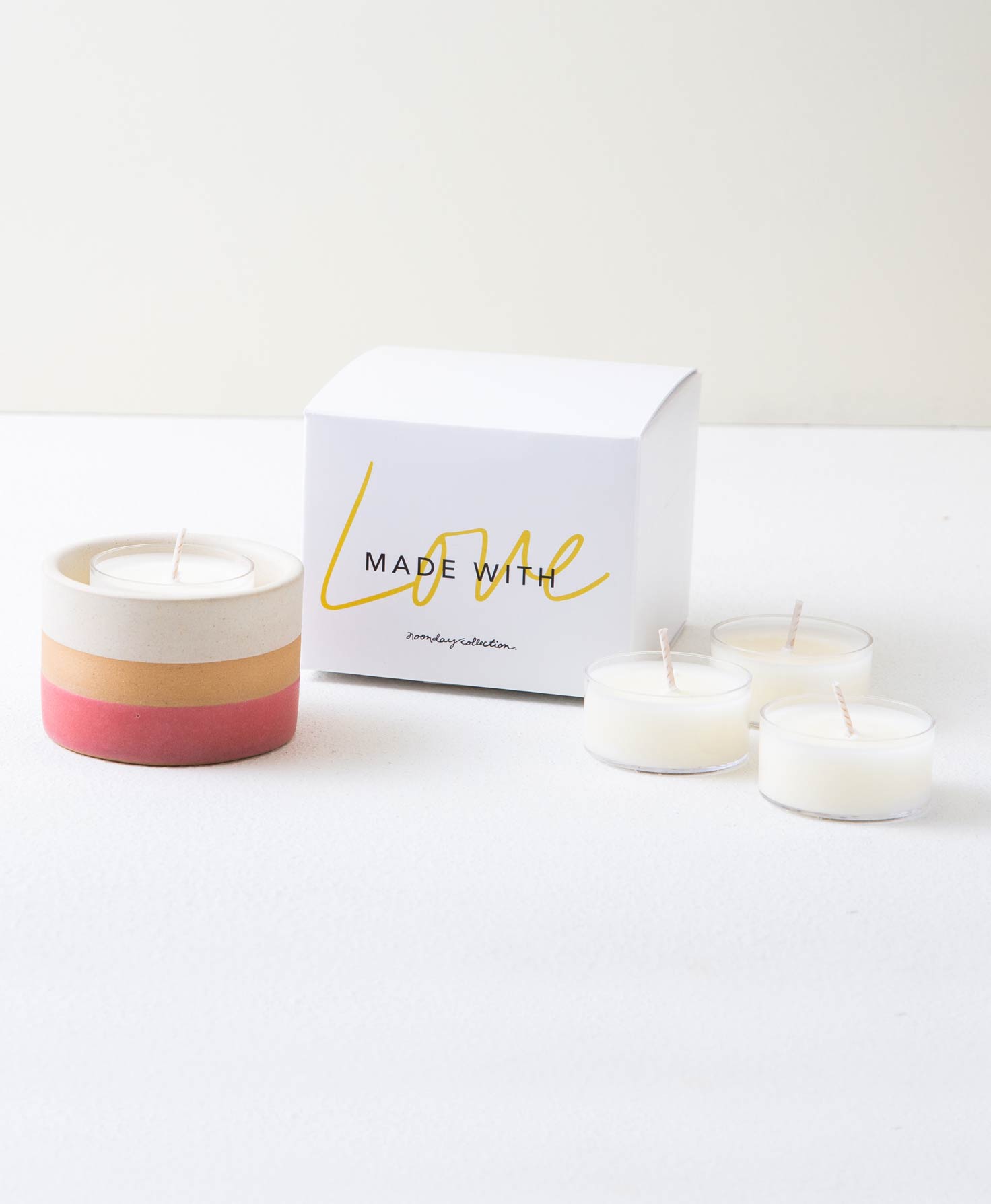 The Banded Stone Tealight Holder in Pink and three white tealight candles sit next to the white box they are packaged in. The box is square and has 